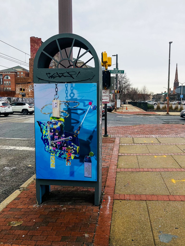 A digital artwork printed on the side of a kiosk is on display on a street.