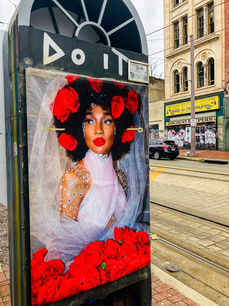 A photograph of a woman with brown skin, adorned with red flowers, on a kiosk displayed outside.
