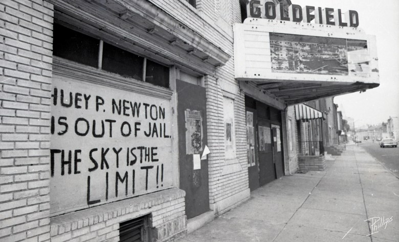 A black and white scan of a derelict theater. A hand painted sign reads: Huey P. Newton Is Out Of Jail. The Sky Is The Limit"
