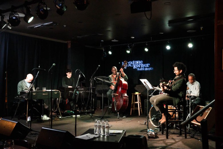 Members of a band sit on a darkened stage with their instruments.