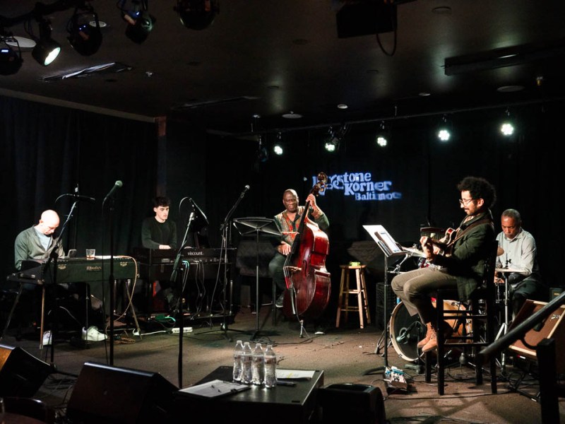 Members of a band sit on a darkened stage with their instruments.