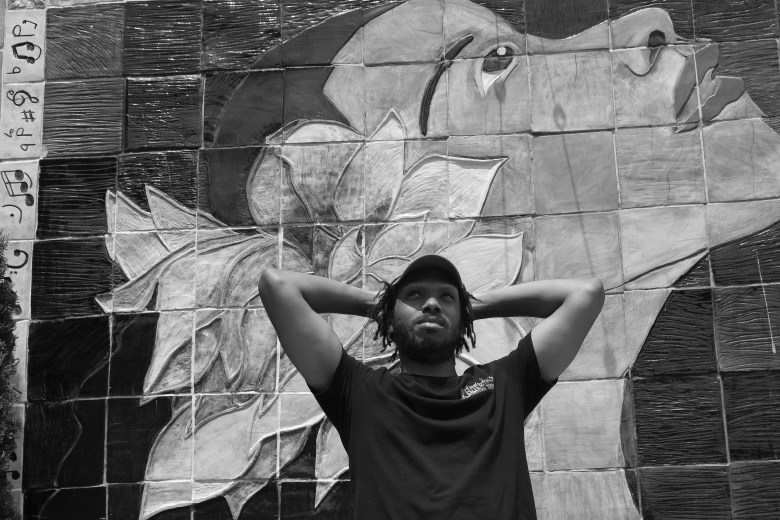 black and white photograph of a man with brown skin posing in front of a mural 