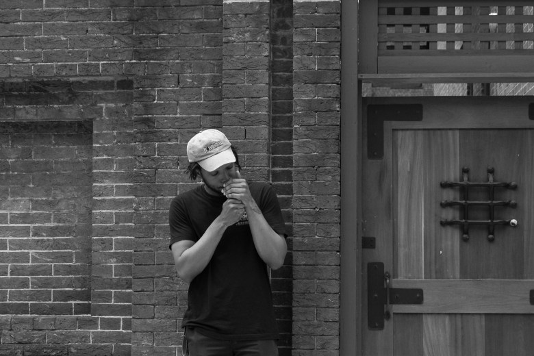 a black and white photograph of a man with brown skin posing with his head down in front of a brick background