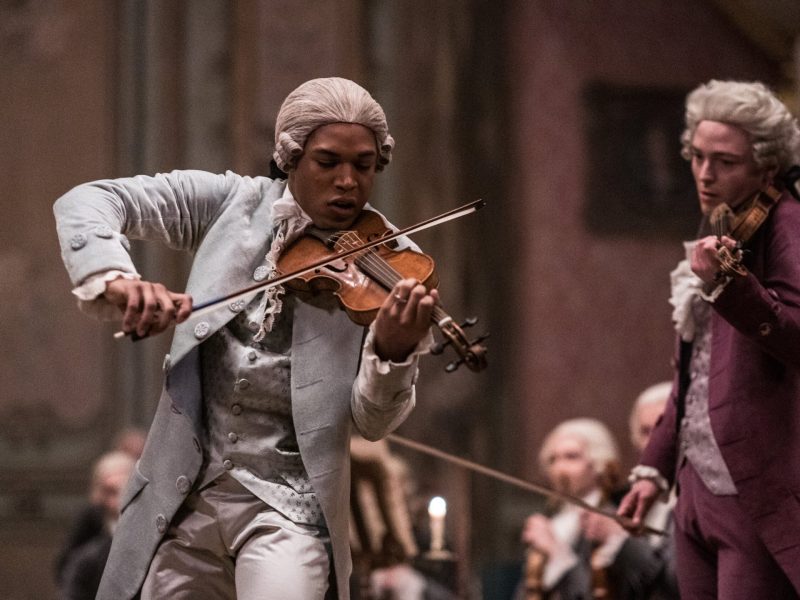 Film still from Chevalier, two men are playing insturmetns in period costumes, the main character has brown skin and a wig and is playing the violin.