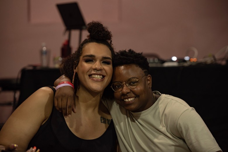 Two people sit smiling facing the camera, they both are smiling and have brown skin.