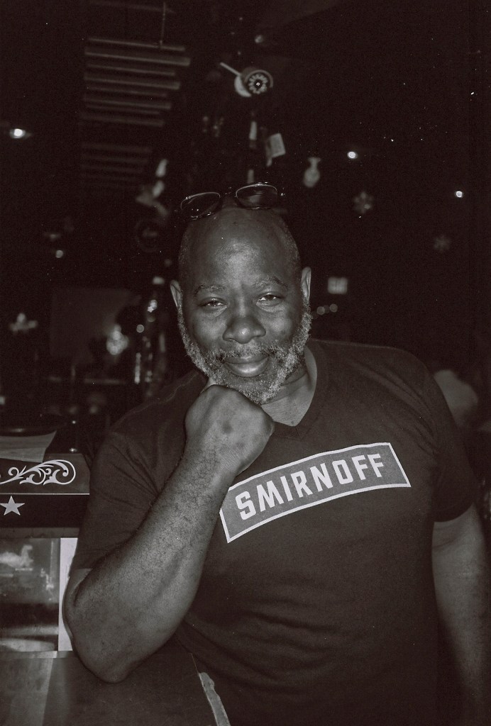 A bearded man poses for the camera. He has a pair of sunglasses on his head and he wears a black t-shirt with "Smirnoff" written on it.