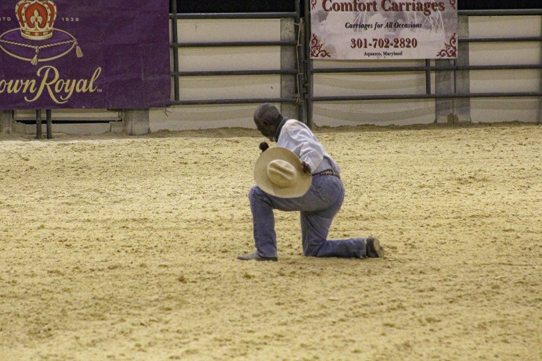 A black cowboy kneels in the center of the arena, he is wearing jeans and holding a tan cowboy hat.