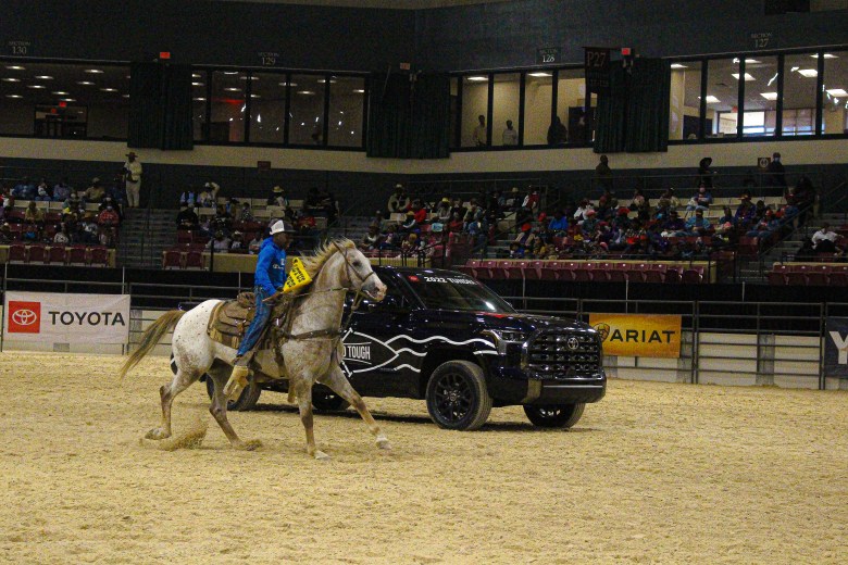 A young cowboy riding in the Bill Pickett Invitational Rodeo, riding a tan horse in the arena alongside a black toyota truck.
