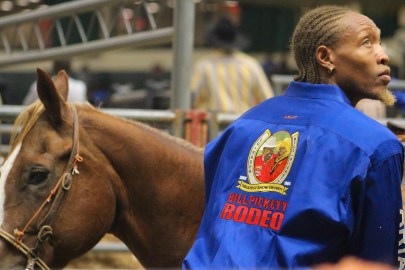 A participant in the 35th Annual Bill Pickett Invitational Rodeo. He is wearing a blue jacket with a Bill Pickett Rodeo logo. There is a horse in the image as well.
