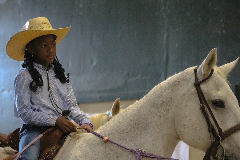 A young competitor in the 35th Annual Bill Pickett Invitational Rodeo. She is a cowgirl with braids and a tan cowboy hat, she wears a blue striped shirt and is riding a white horse.