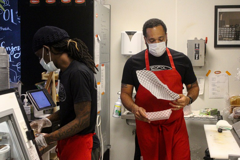 A photograph of two men preparing orders at Neopol Savory Smokery. One has long black hair, brown skin, and tattoos. The other has short brown hair and brown skin. They both are wearing back t-shirts and red aprons.