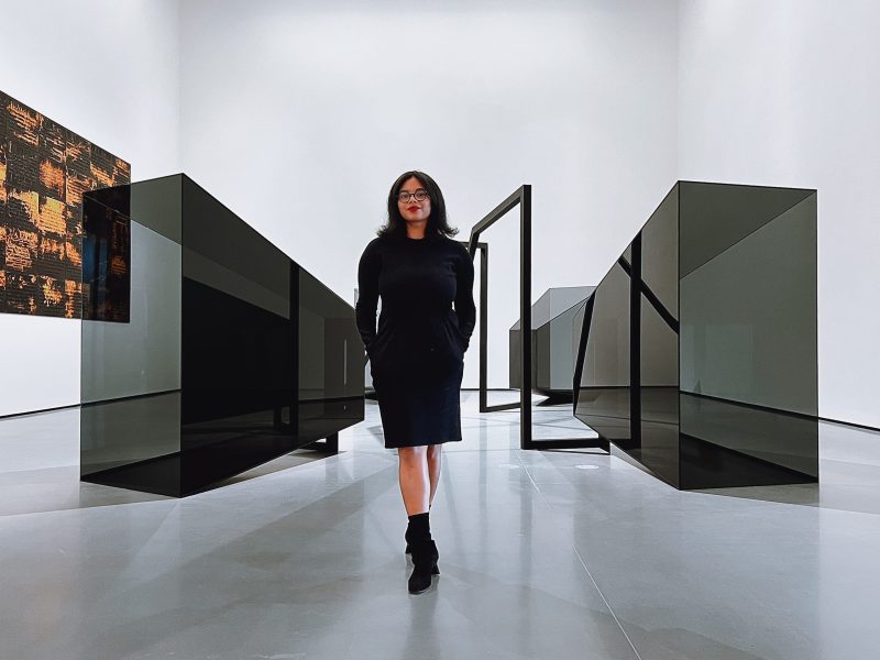 A woman with brown skin stands in a gallery in front of an artwork.