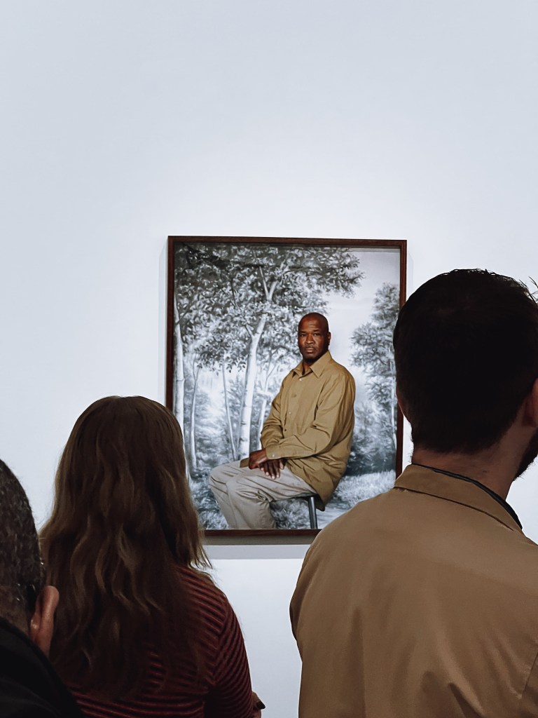 Three visitors view an artwork. It is a photo of a Black man seated. Wearing a tan shirt and tan pants.