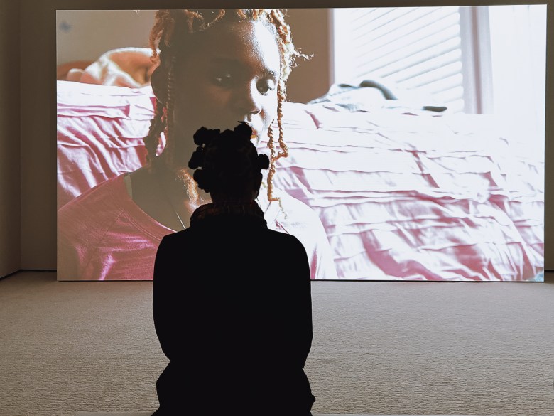 A woman is seated in front of a large video projection. She is facing the screen.