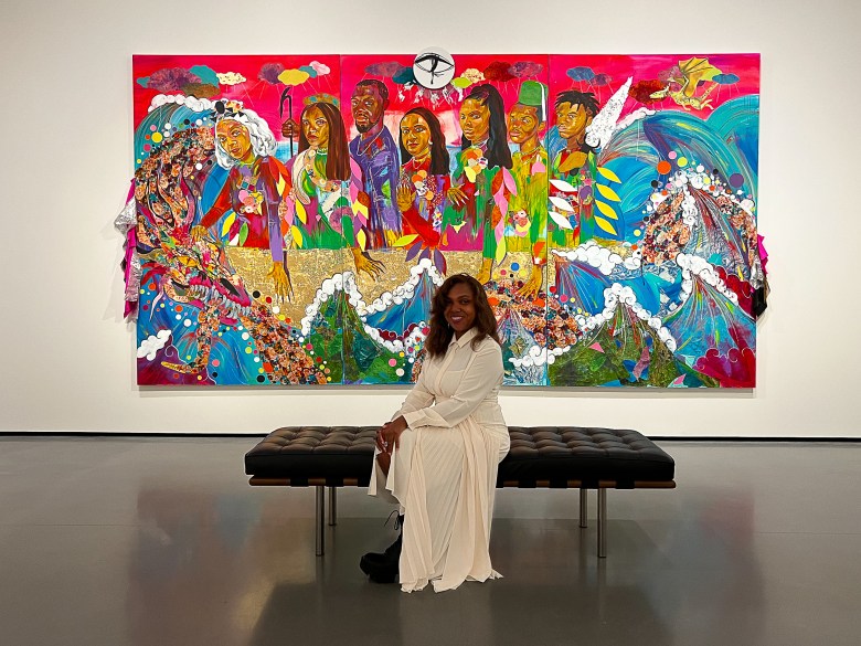 A Black woman is seated in front of a large scaled artwork, she is wearing a white dress and is smiling.