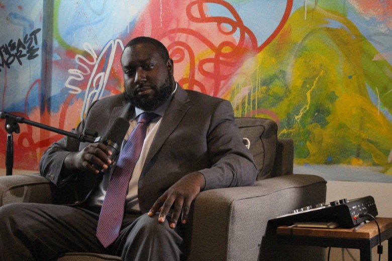A man with brown skin is seated in front of an artwork, wearing a suit and holding a microphone. 