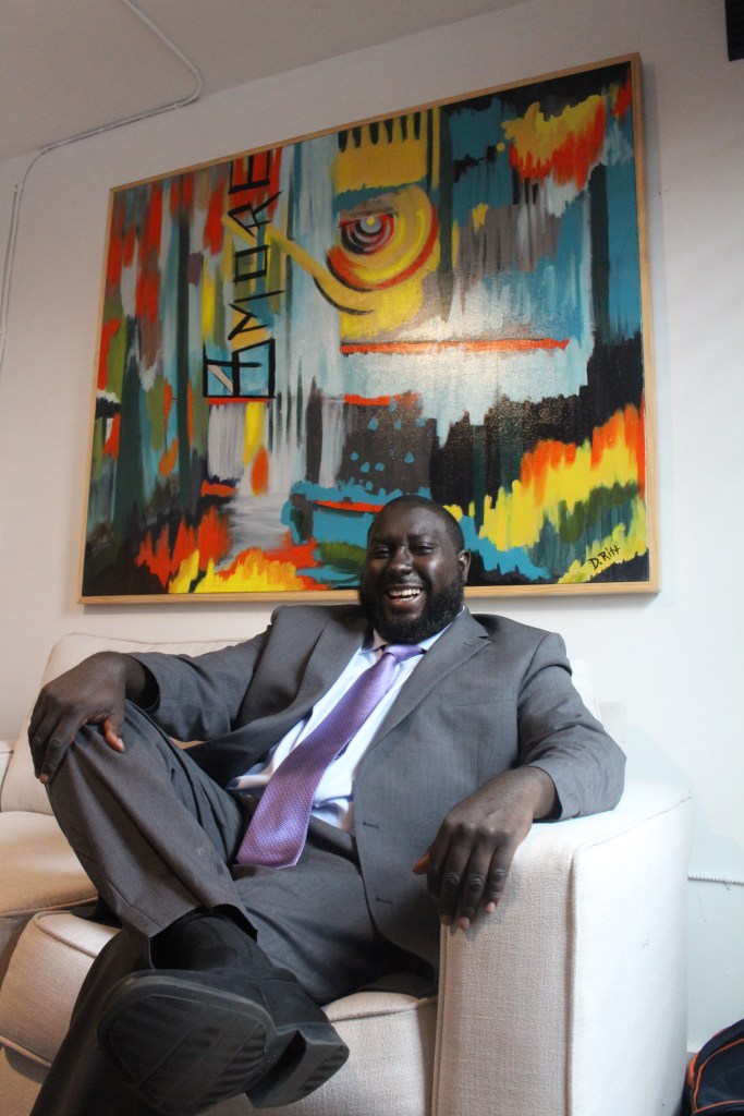 A man with brown skin wearing a suit is seated and smiling in front of a multicolored artwork. 