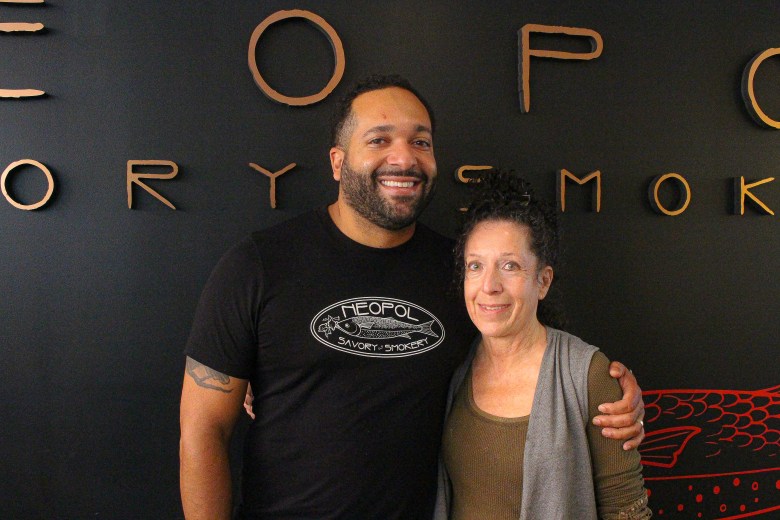A man and a woman smile in front of a black wall. He is wearing a black tshirt that says neopol savory smokery, she is wearing an olive green t-shirt and a grey vest.