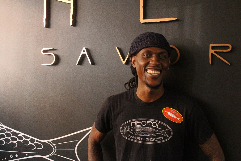 A man with brown skin smile at the camera in front of a black wall. He has dreadlocks and is wearing a shirt that says Neopol Savory Smokery.