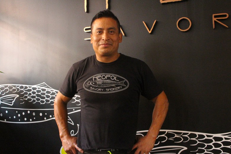 A man with brown skin and short hair smiles at the camera. He is wearing a Black Neopol savory smokery T-shirt.