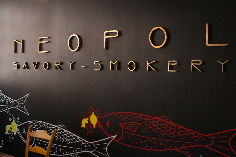 A wall painted black with gold letters that read NEOPOL SAVORY-SMOKERY. There is a painted mural of fish at the bottom of the wall.