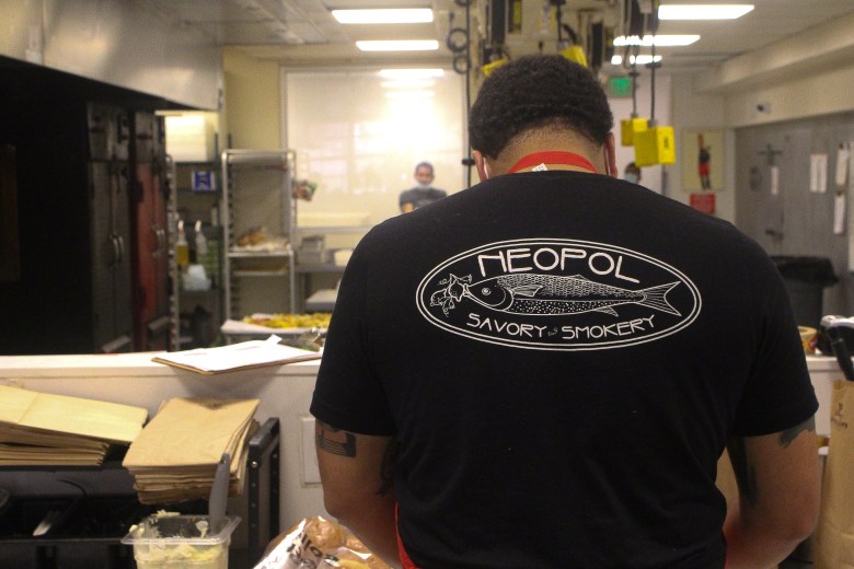 A man prepares a food order with his back turned to the camera. He is wearing a black t-shirt that says Neopol Savory Smokery in white letters.