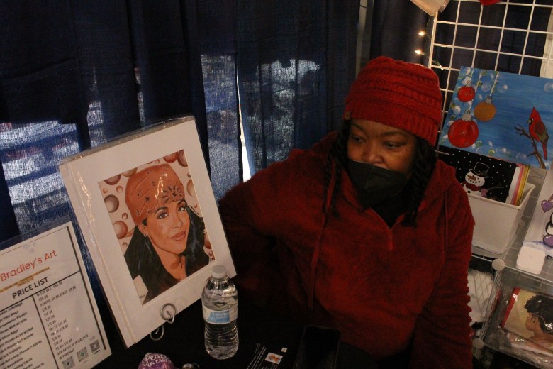 A woman with brown skin wearing a mask seated next to her artwork. 