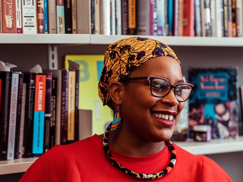 A photograph of Jessica Marie Johnson, associate professor, Department of History, Johns Hopkins University. She is a woman with brown skin, wearing a head scarf, glasses, a red top and a necklace