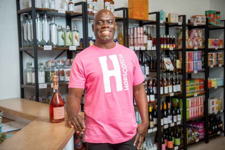 A man smiles next to a bottle of spirit free rose, he has brown skin and is wearing a pink t-shirt. 