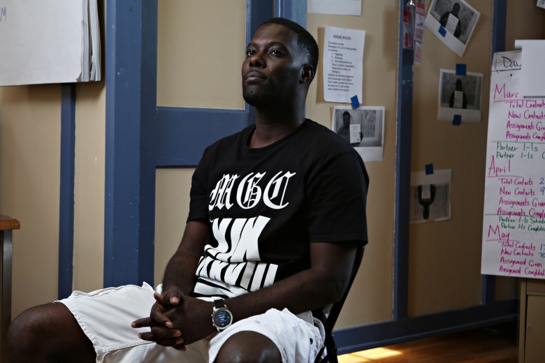 A photo of a Black man sitting in a chair. He is wearing a black t-shirt with white words on it and white shorts.