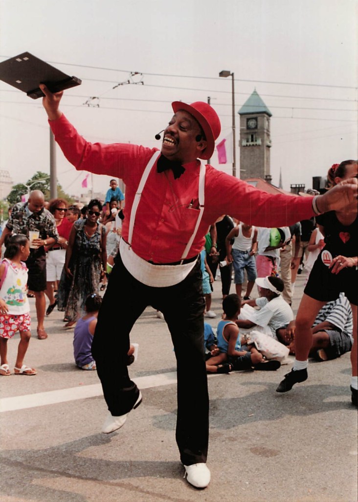 Children's television star Kinderman dances and sings for the crowd. He wearts a red hat, red shirt, black bow-tie, and white suspenders. He also has on black pants and white shoes.