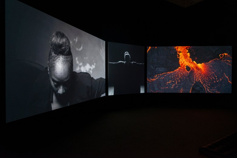 Three screens in a dark room. One shows the top of a person's head. One shows a person with their arms outstretched, and one shows a fiery, glowing substance.