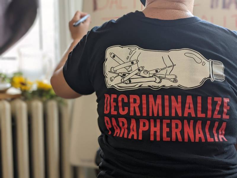 A photo of someone's back. They are wearing a black t-shirt that reads "decriminalize paraphernalia" in red letters.