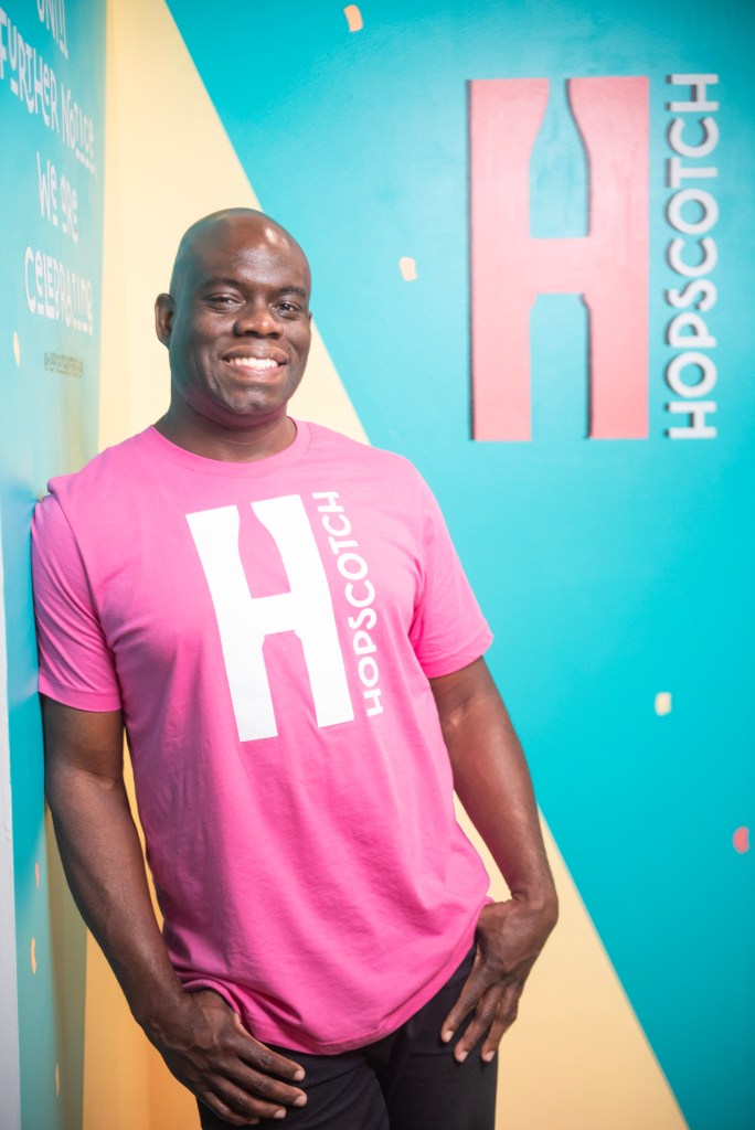 A man with brown skin wearing a pink shirt smiles in front of a multicolored wall. 