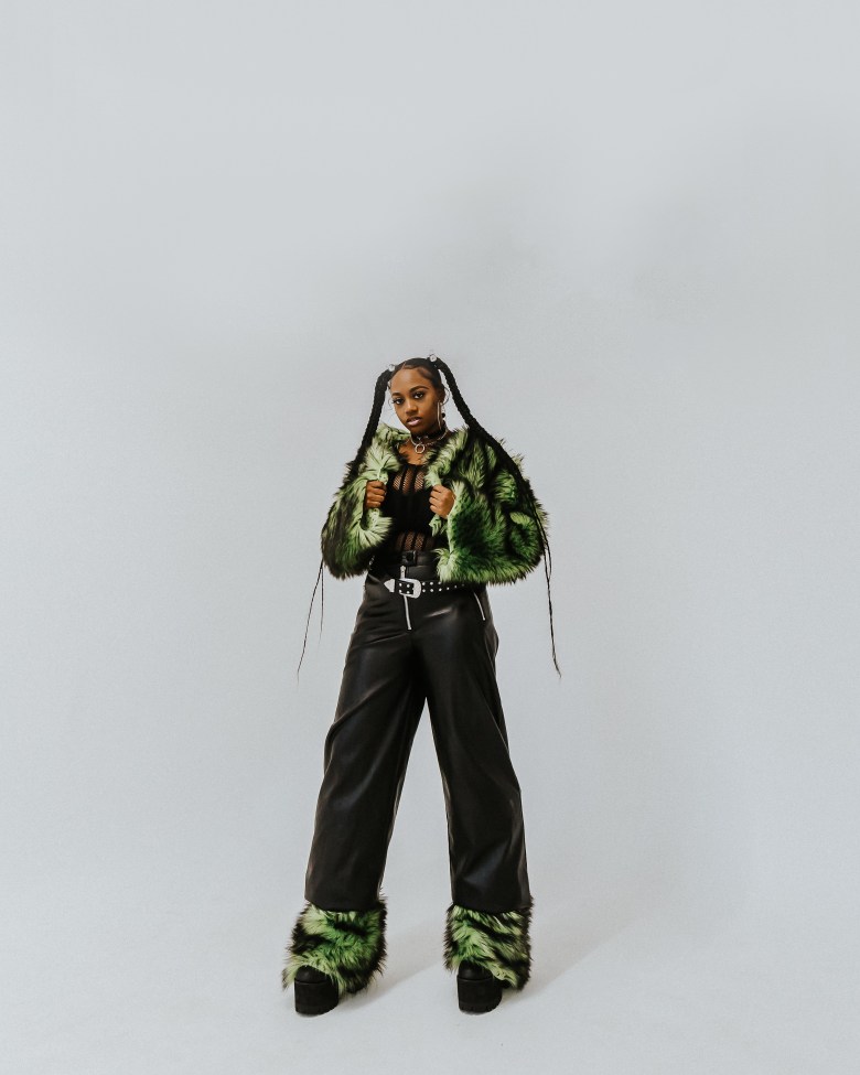 A woman with brown skin poses. She has on a green jacket and dark pants.
