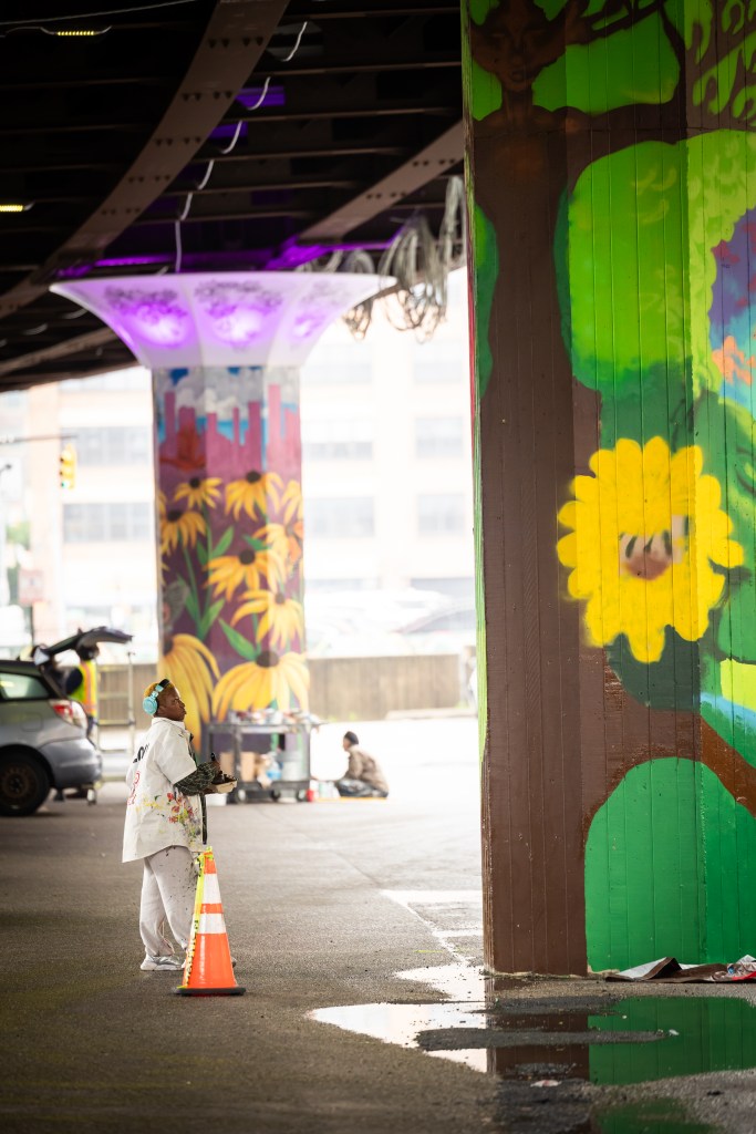 Artist Afr0delic looks up at a pillar they are painting under the Jones Falls Expressway.