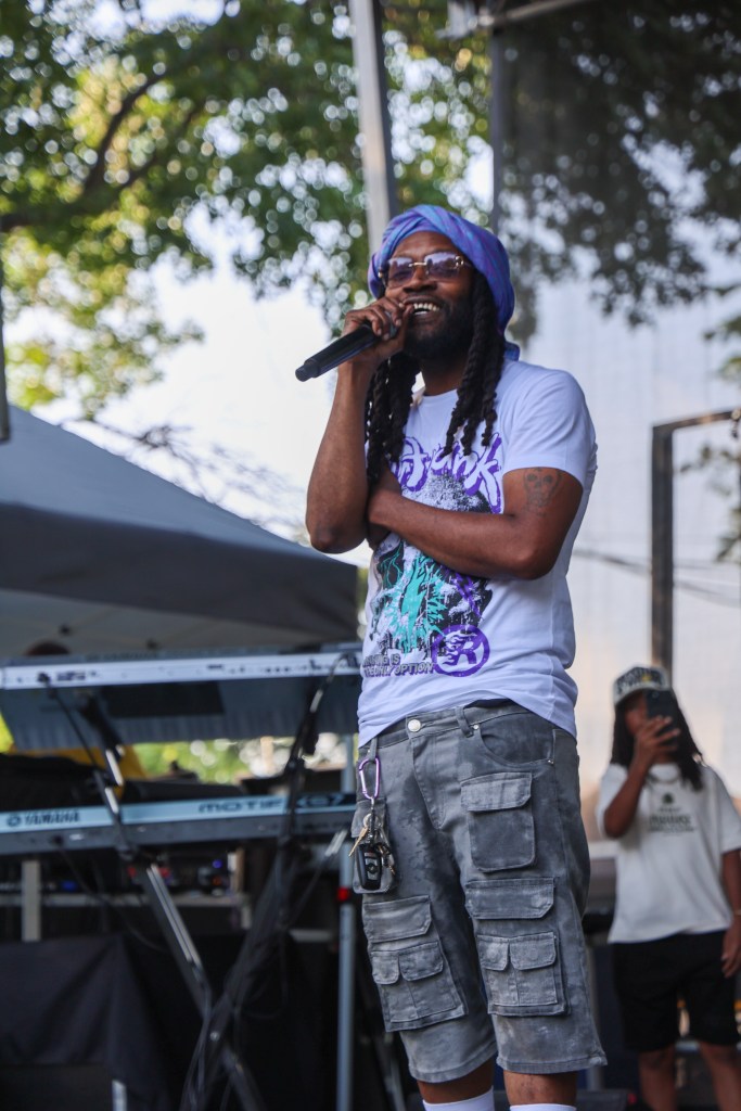 A Black man with long locs smiles as he holds a microphone. He has on a purple head wrap, a purple t-shirt, and cargo shorts with a camouflage print.