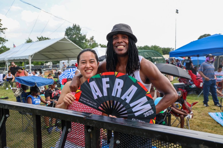 A photo of two people smiling. One person has long locs and wears a hat. The other holds a fan that has the words "AFRAM 2025" on it.