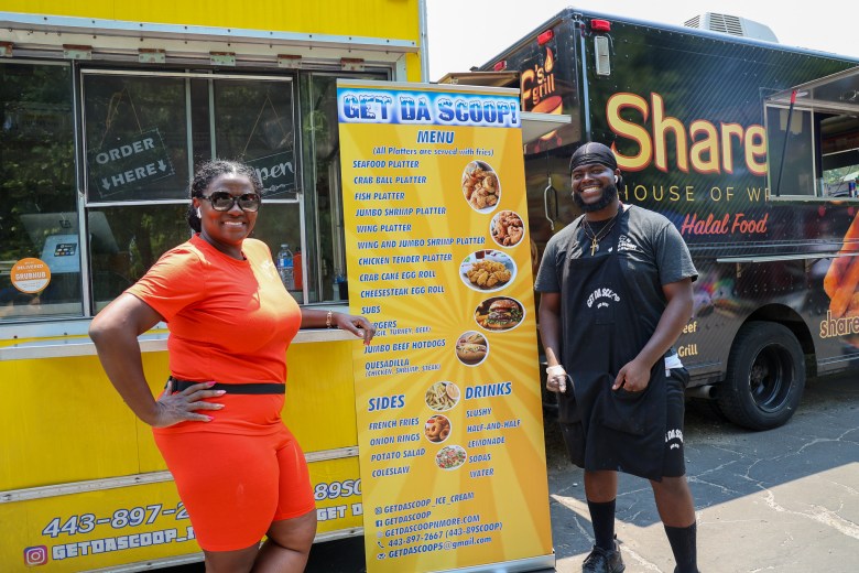 two people pose by a sign near a food truck