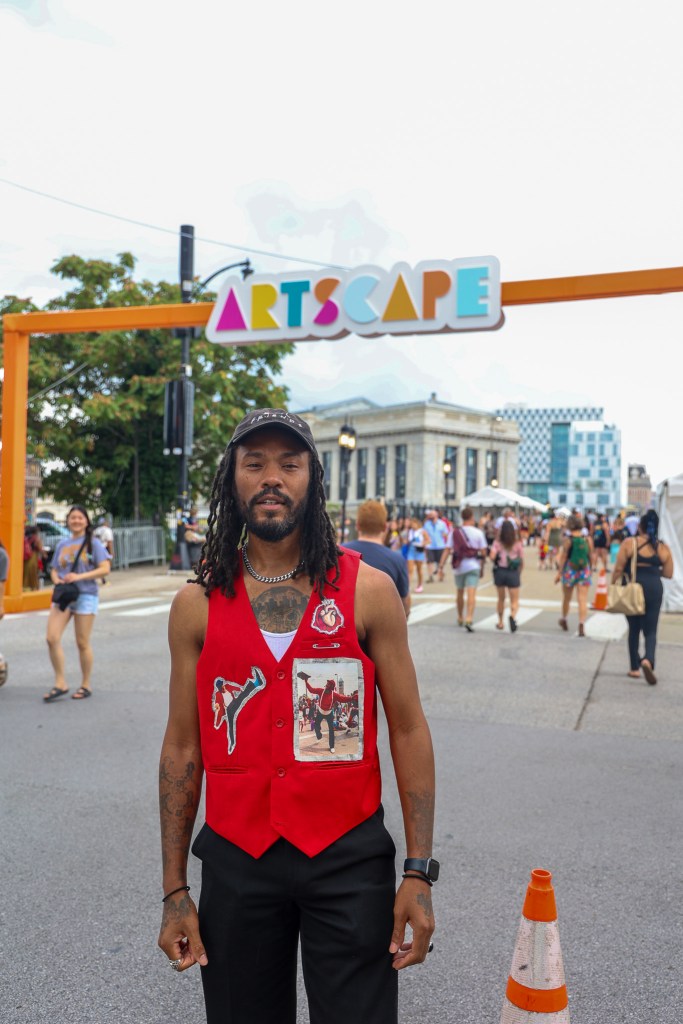 A person wearing a red vest stands in front of a sign that says artscape