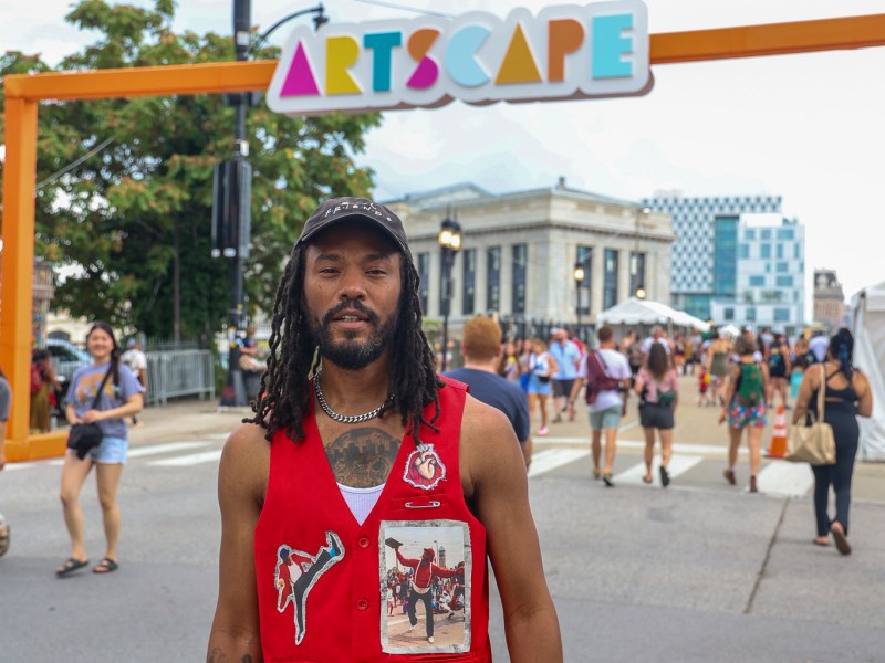 A person wearing a red vest stands in front of a sign that says artscape