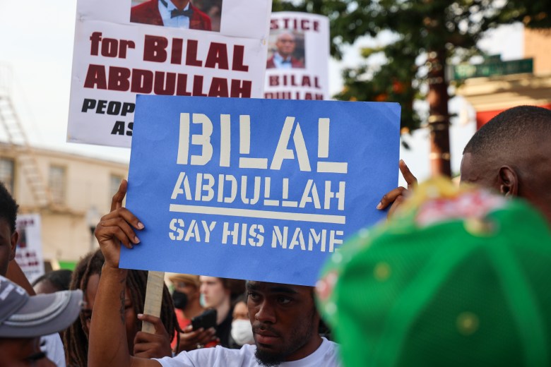 A person holds a blue sign. In white are the words "Bilal Abdullah say his name." Other people holding signs can be seen in the background.
