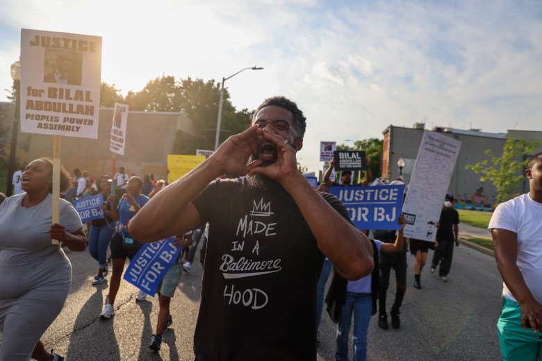 Aaron Maybin, a Black man, chants at a march. He wears a black t-shirt that says "made in a Baltimore hood." Othe people march with him.