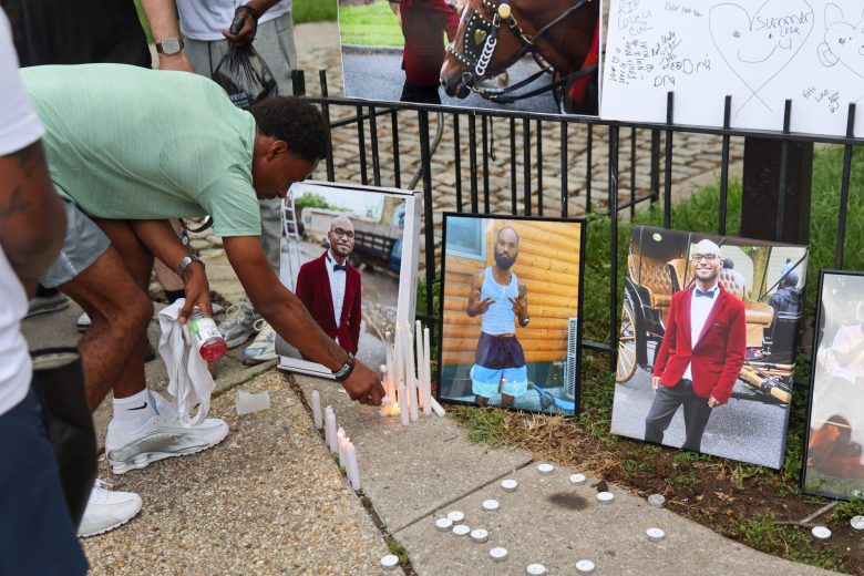 A Black person wearing a green shirt and gray shorts crouches down to light a white candle. On the ground are white candles scattered around and photos of a Black man.