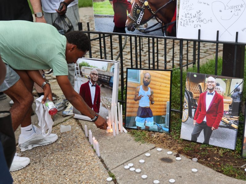 A Black person wearing a green shirt and gray shorts crouches down to light a white candle. On the ground are white candles scattered around and photos of a Black man.