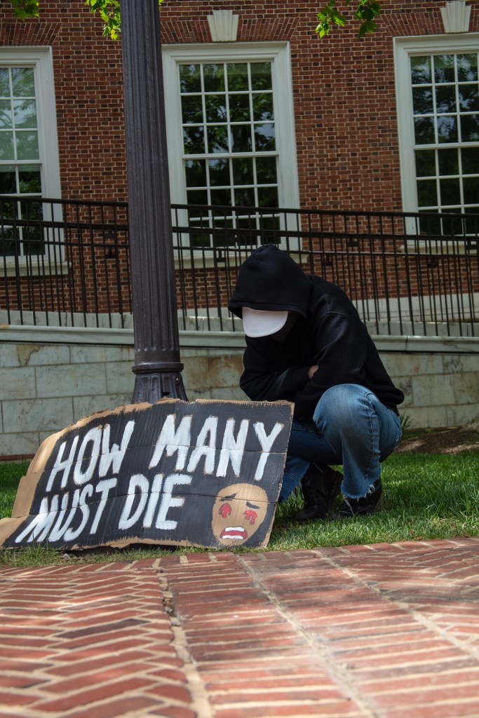 A person wearing a black hoodie crouches near a sign that reads: "how many must die."