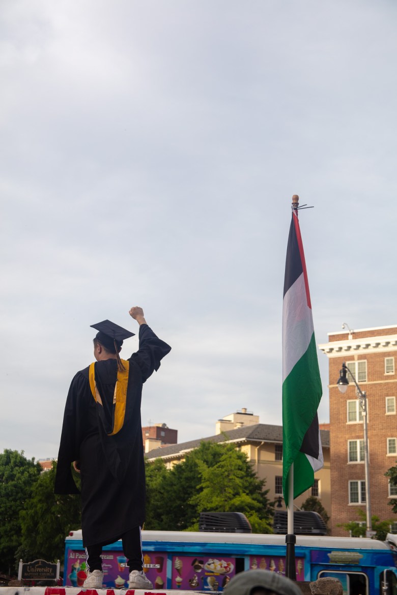 A person dressed in a graduation cap and gown raises their fist as they stand next to the flag of Palestine.