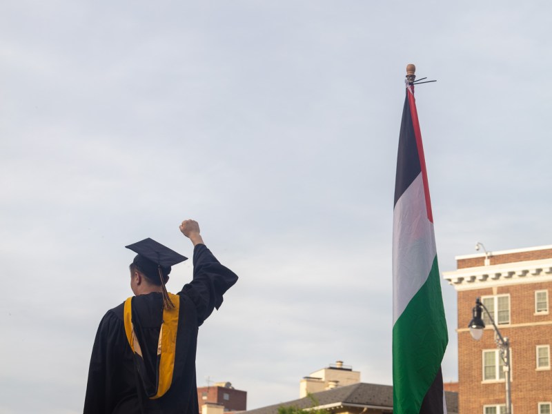 A person dressed in a graduation cap and gown raises their fist as they stand next to the flag of Palestine.