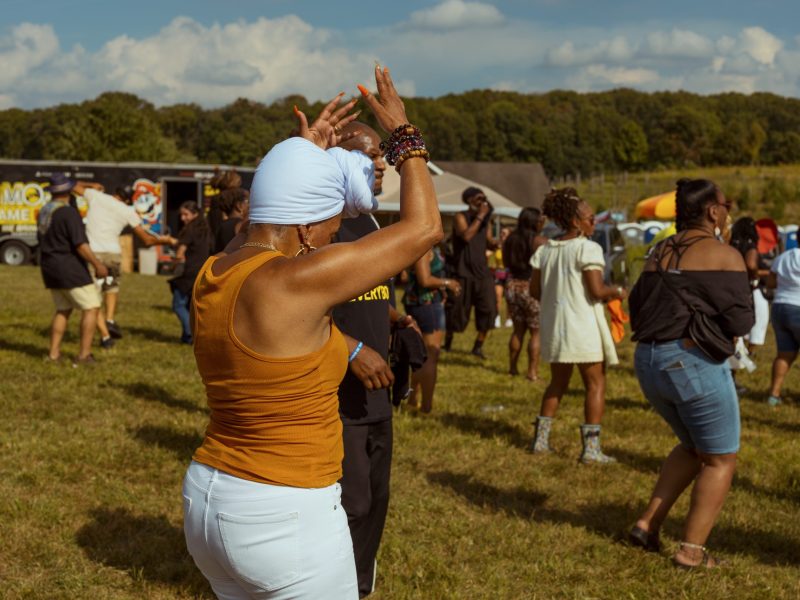 A woman in a white head wrap dances at the Collective Minds outdoor music festival. She is a Black woman wearing an orange top and white pants