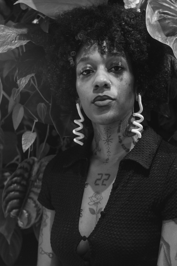 Black and white photograph portrait of a woman with brown skin, she is wearing white sculptural earrings, a black knit top, and has several tattoos. She looks at the camera and is photographed in front of leaves.
